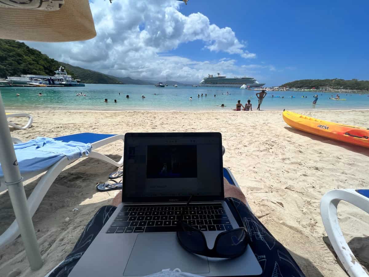 Alan Huthison working on his computer on the beach at Labadee, Haiti with Vision of the Seas cruise ship in the background